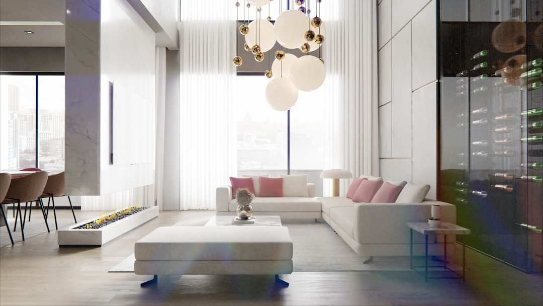 This image showcases a stunning, contemporary open-plan living and dining space defined by its soaring ceilings and abundant natural light. A plush, white sectional sofa, accented with soft pink cushions, invites relaxation, arranged around a matching ottoman and low coffee table. It faces a sleek, modern fireplace set into a low console. Dominating the vertical space, a dramatic cluster pendant light with large white globes hangs from the high ceiling, adding a sculptural element. To the right, a floor-to-ceiling glass wine cellar reveals an impressive collection, reflecting the rooms light